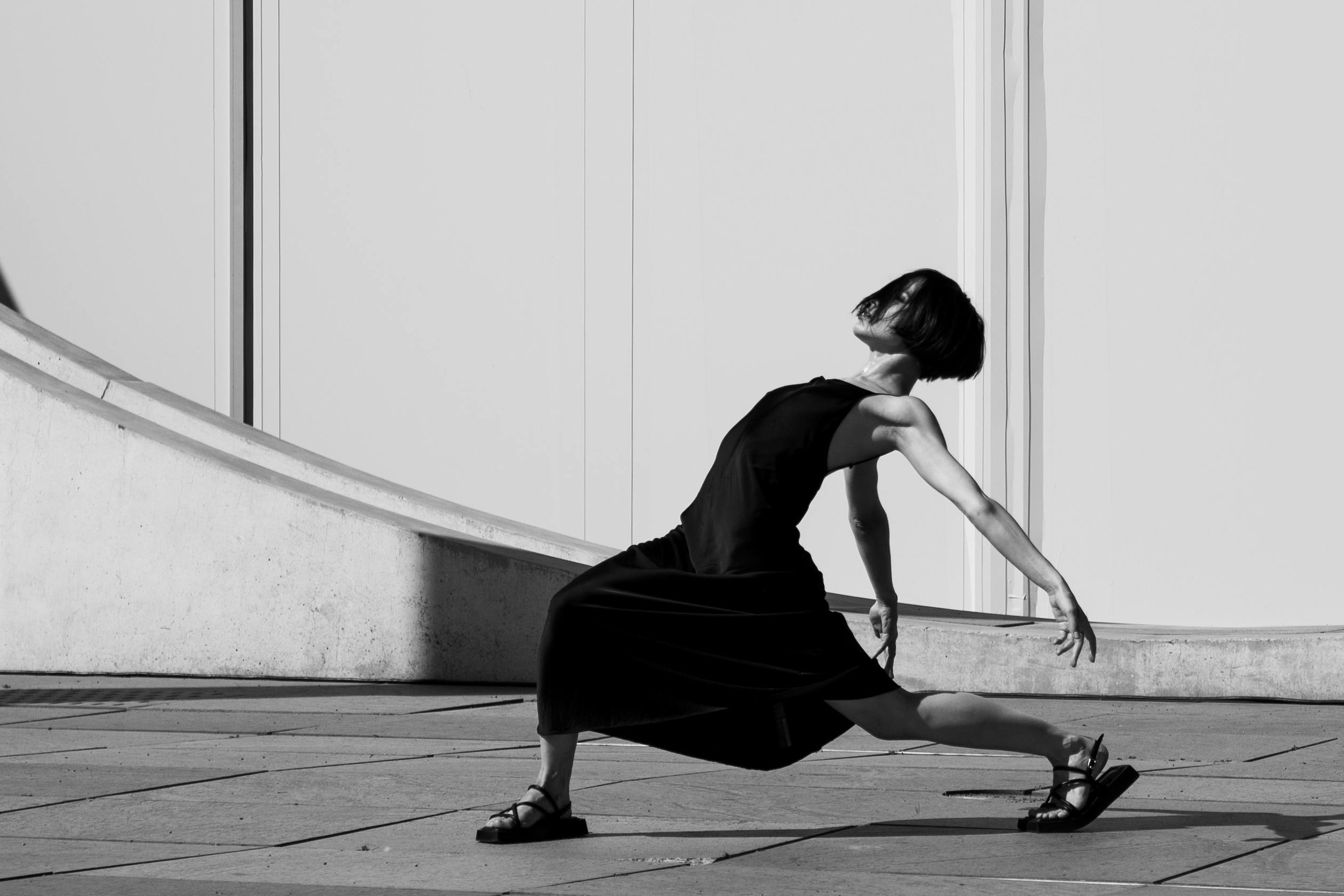 A person with short hair wearing a sleeveless black dress and sandals performing a dance move outdoors on a tiled surface, with a blank wall and a sliding door in the background, in black and white.