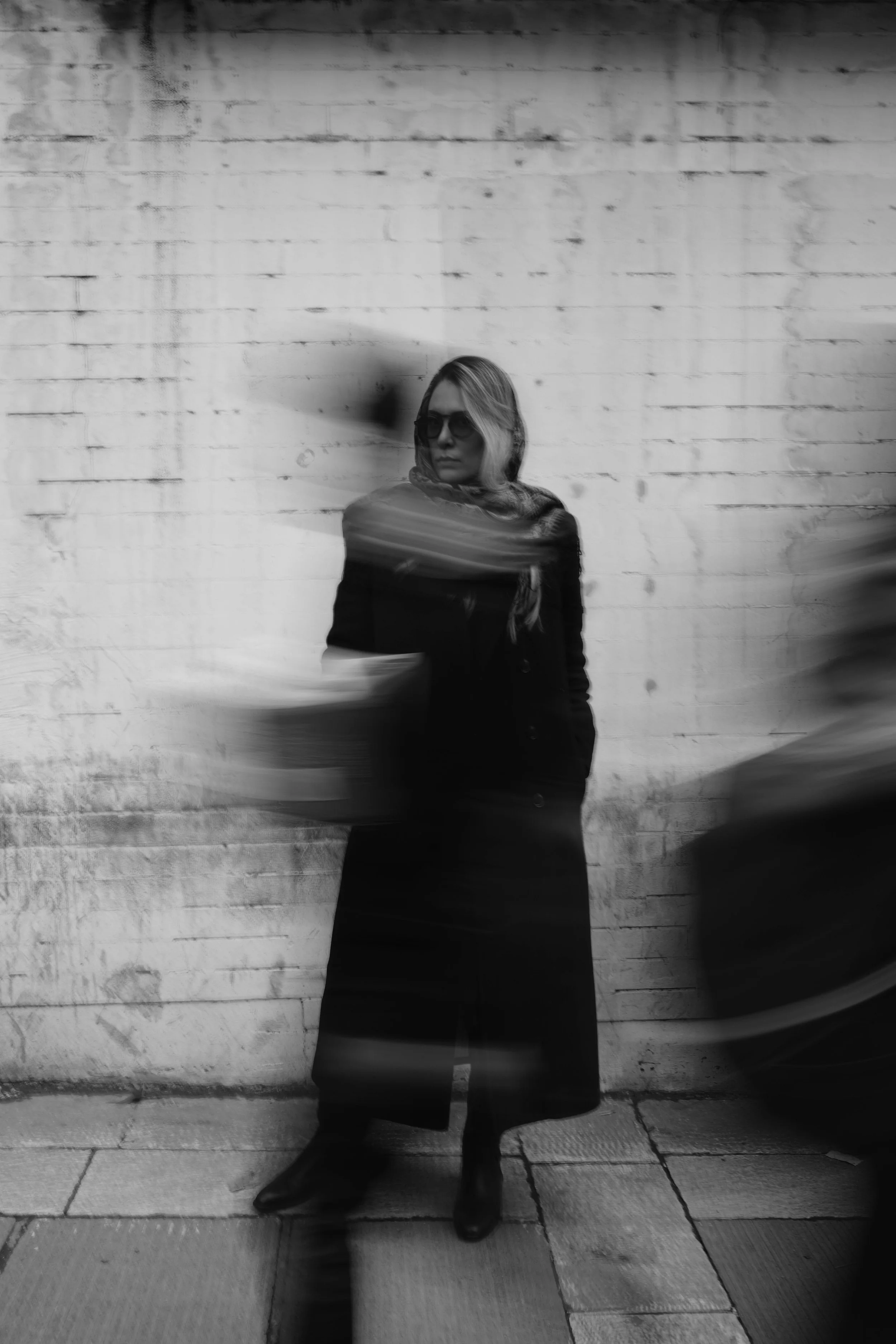 A woman wearing sunglasses and a long coat stands against a white brick wall, with blurred figures passing by in motion.
