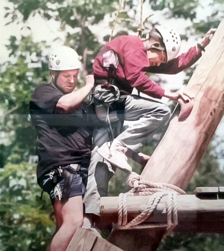 Two individuals climbing a wooden structure, both wearing helmets and harnesses for safety.
