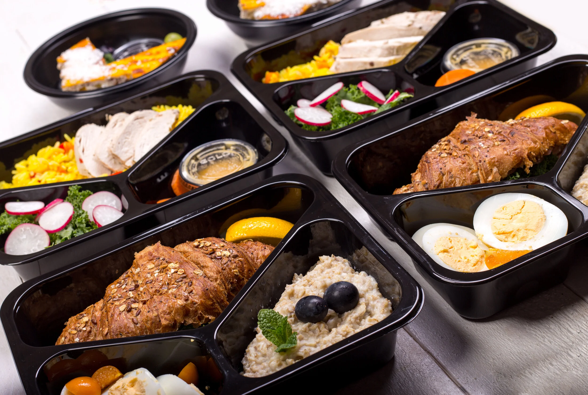 Multiple black meal prep containers filled with various foods, including sliced turkey, rice, salad with radishes, roasted chicken, hard-boiled eggs, mashed potatoes with blueberries, hummus, and lemon slices.