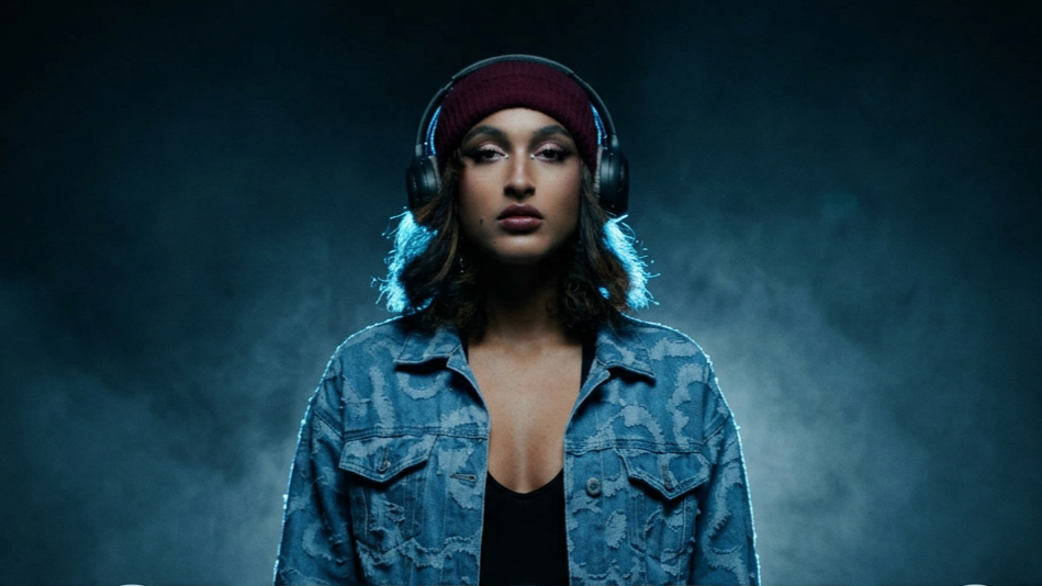 A young woman wearing a maroon beanie and large black headphones, with shoulder-length wavy dark hair, is standing against a dark background, looking directly at the camera with a neutral expression, dressed in a denim jacket over a black top.