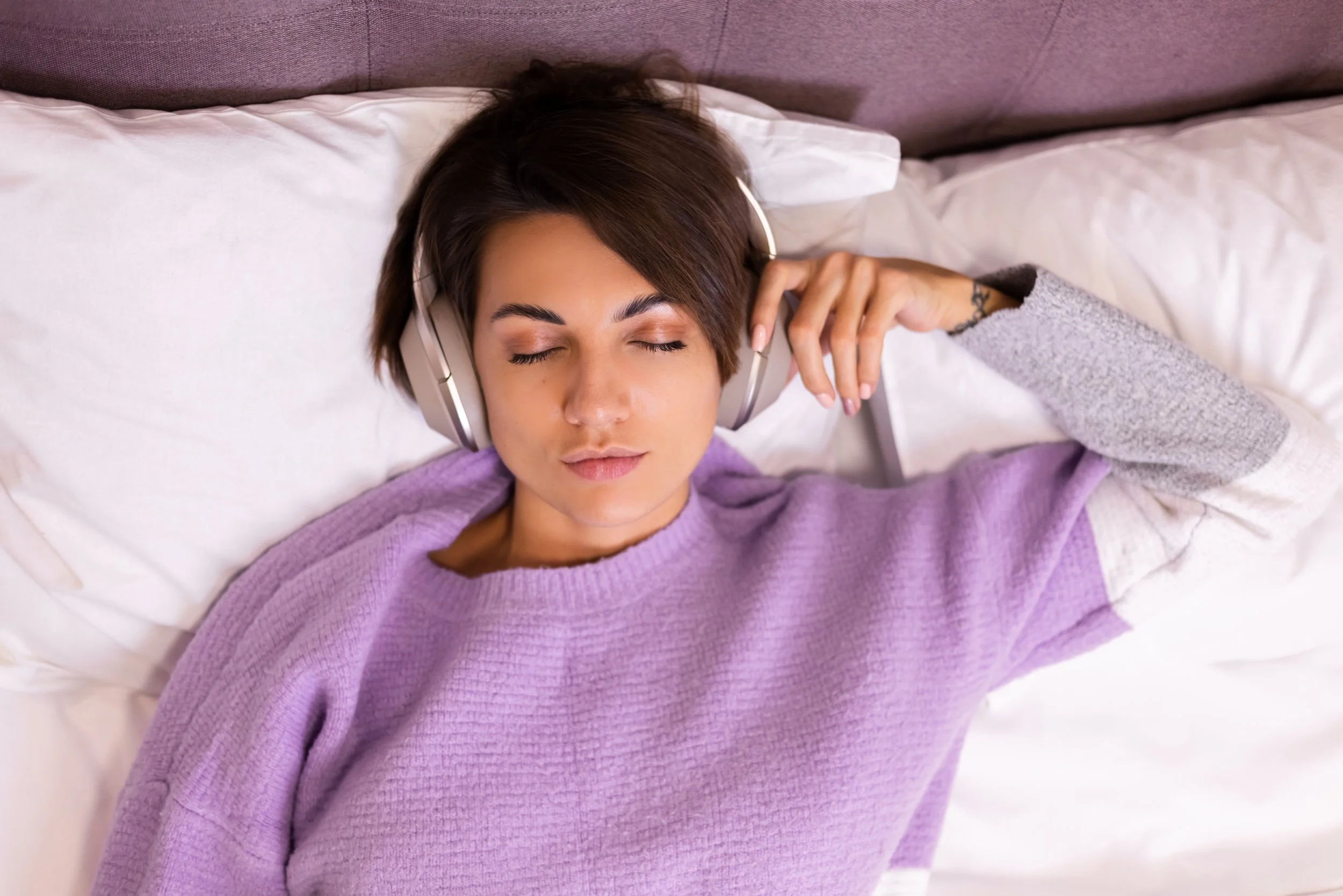 Woman lying in bed wearing headphones and a cozy sweater with eyes closed, listening to something calming