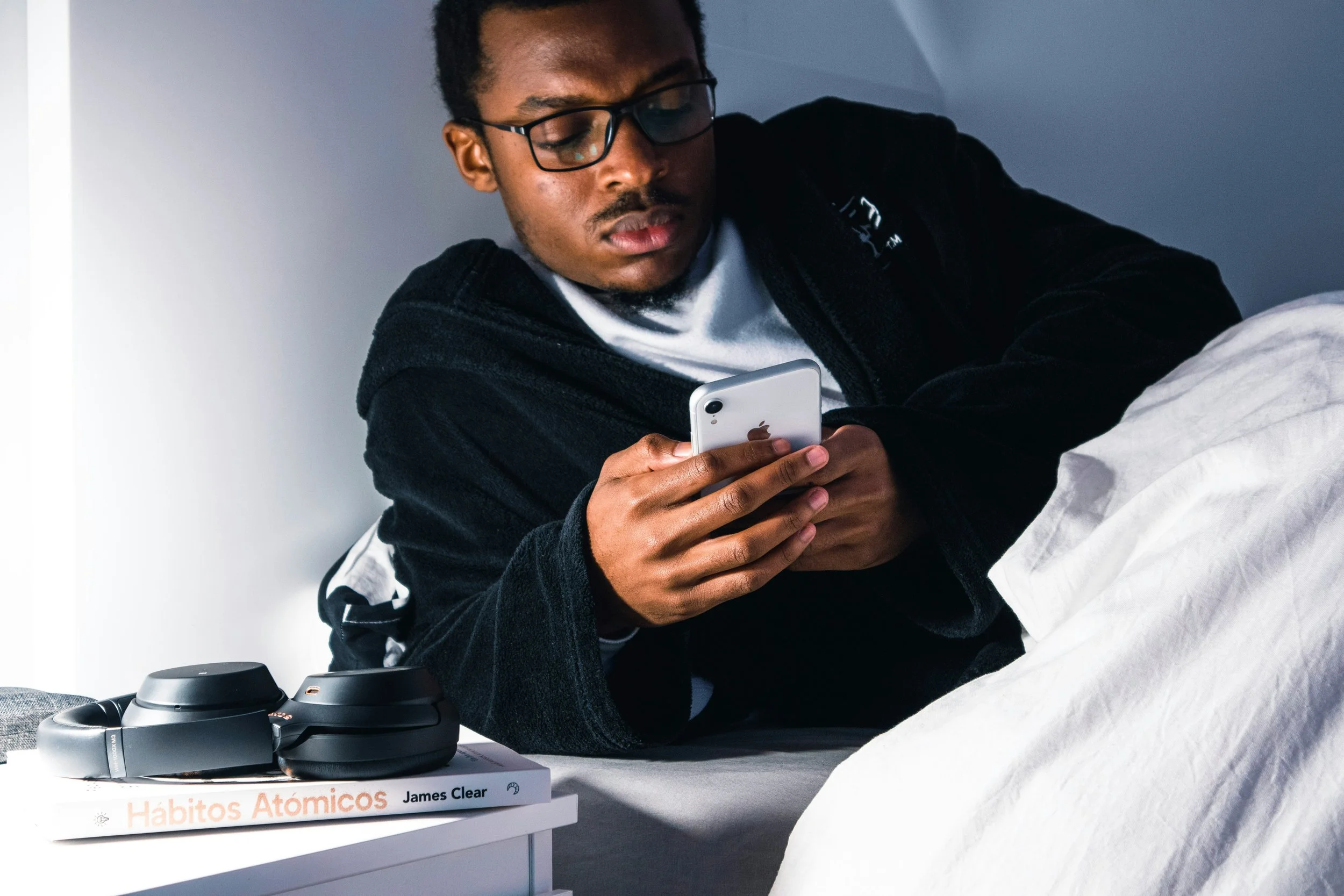 Man lying in bed at night scrolling through his phone, with headphones and a book on the nightstand beside him