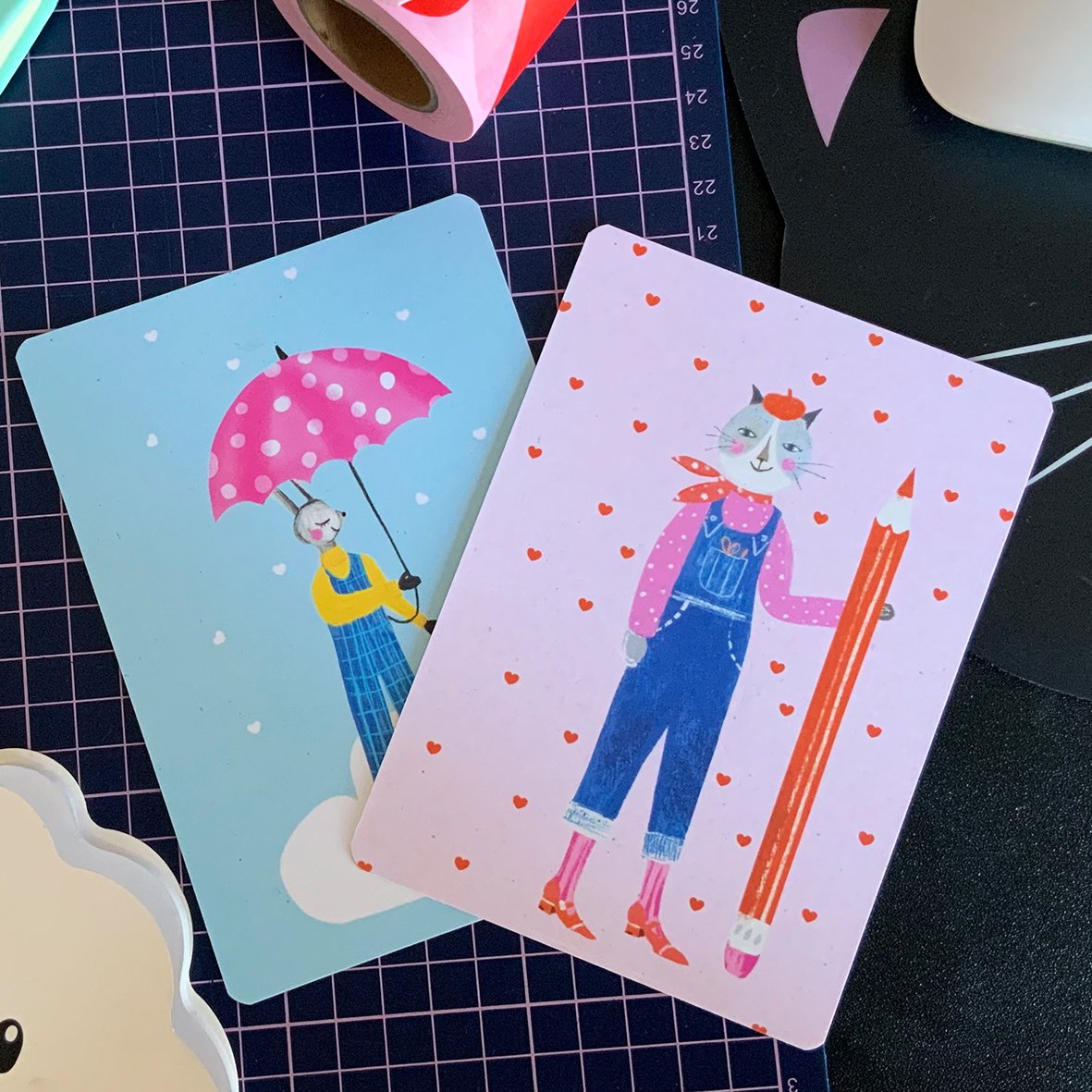Illustrations of anthropomorphic animals, one of a rabbit holding a pink polka-dotted umbrella in the snow, and a cat dressed in overalls, holding a large pencil, surrounded by small red hearts.