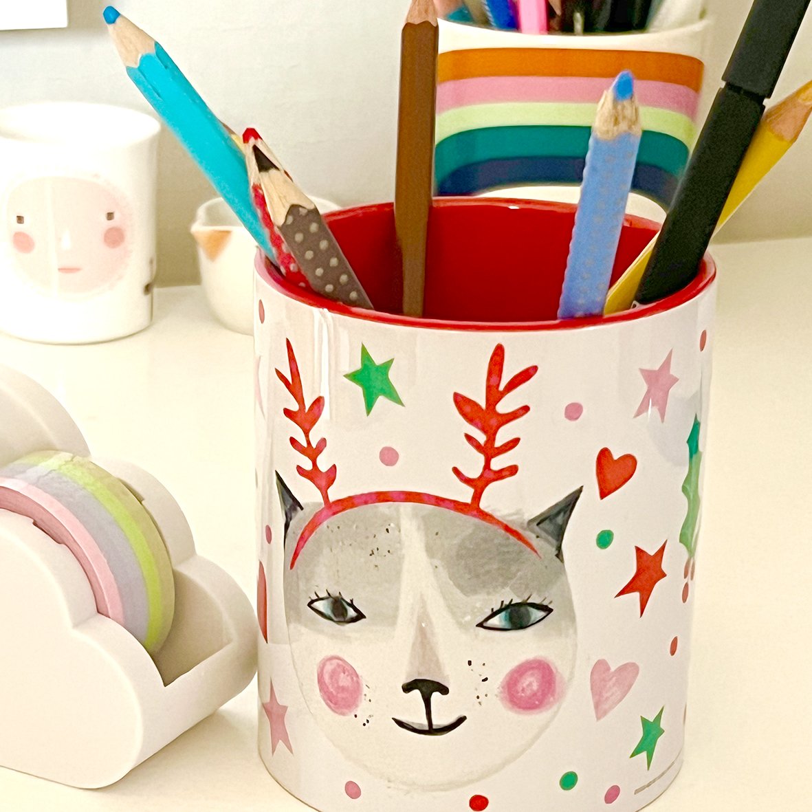 A decorated can featuring a cat face with closed eyes, blush on cheeks, and surrounded by colorful stars, hearts, and antlers, used as a holder for colored pencils.