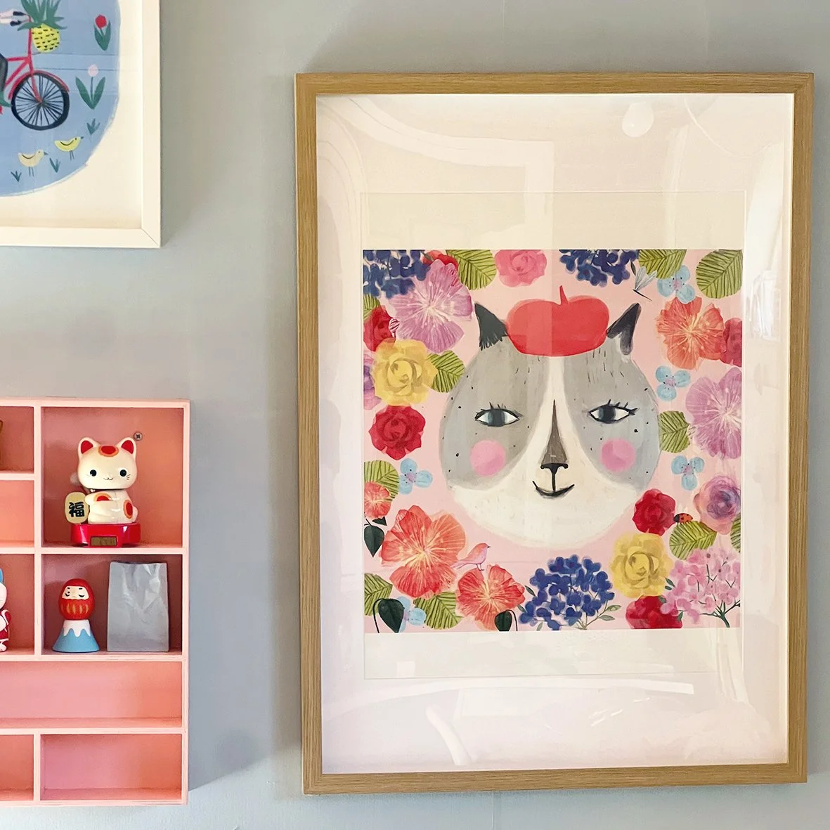 Decorative wall art of a cat's face surrounded by colorful flowers, framed in wood, with a pink shelf holding a lucky cat figurine and other items nearby.