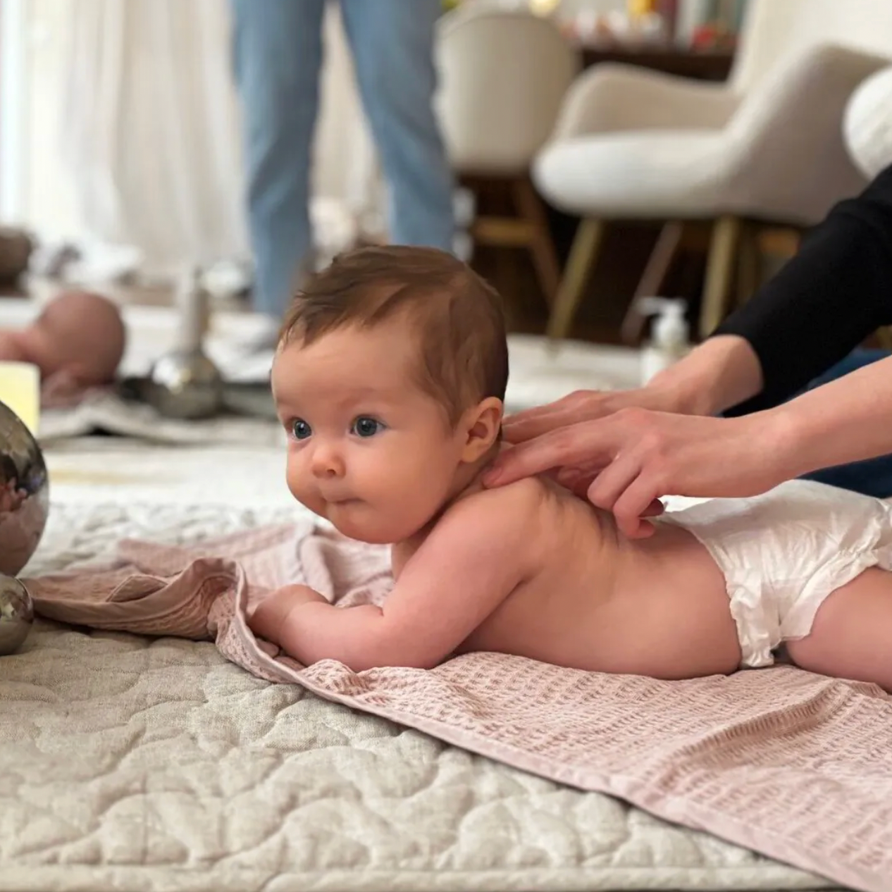 A baby lying on a quilted blanket, getting a massage from an adult, with a blurred background of furniture and a person in jeans.