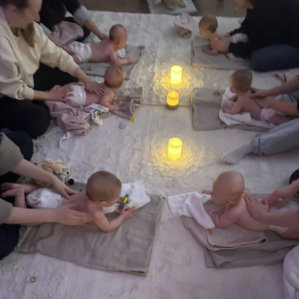 A group of newborn babies lying on towels and blankets on the floor, being gently held and massaged by adults, with yellow candles in the center providing soft lighting.