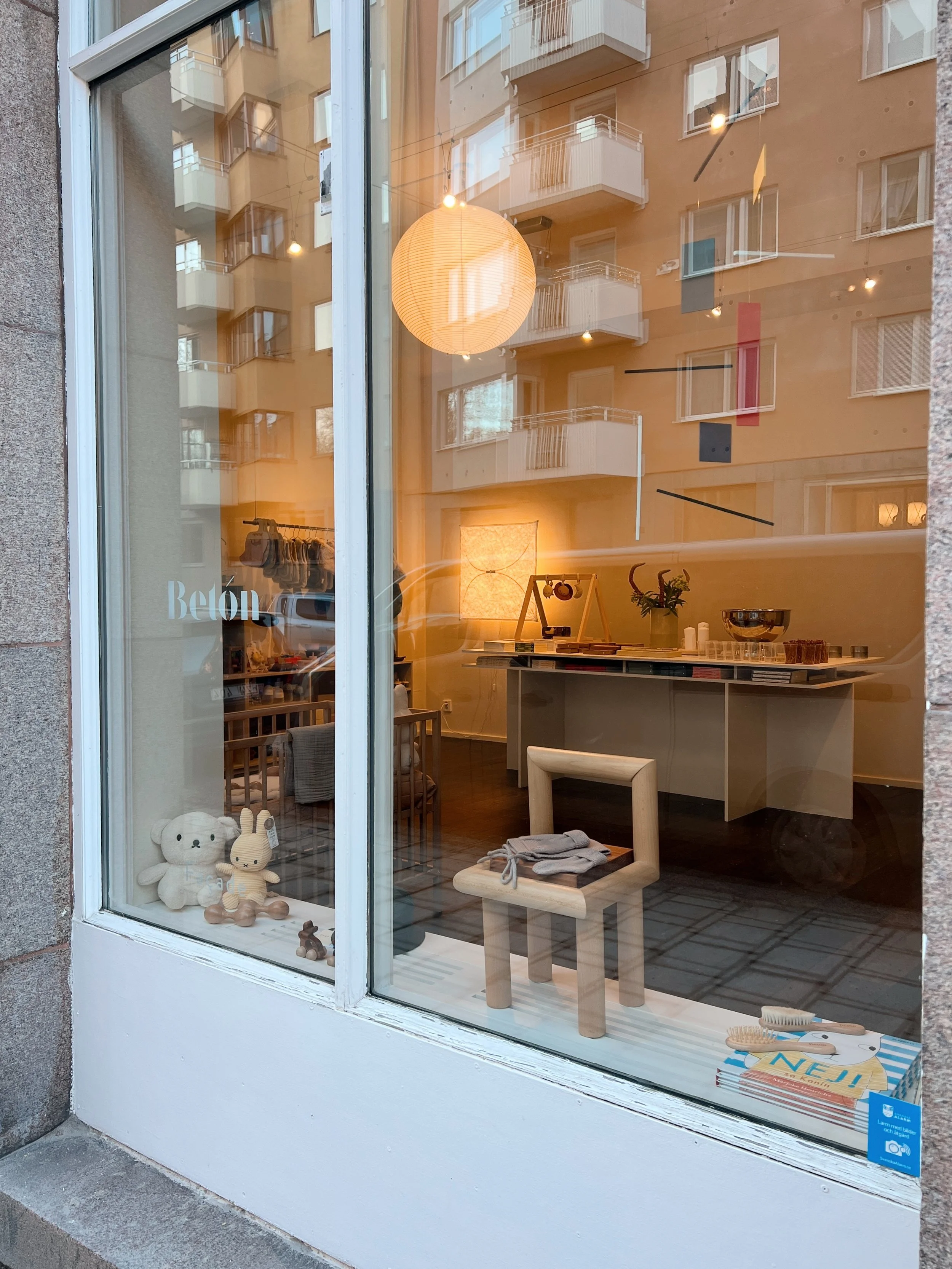 View through a shop window displaying children's toys and furniture, including stuffed animals, a small chair, and hairbrushes, with apartment buildings reflected in the glass.