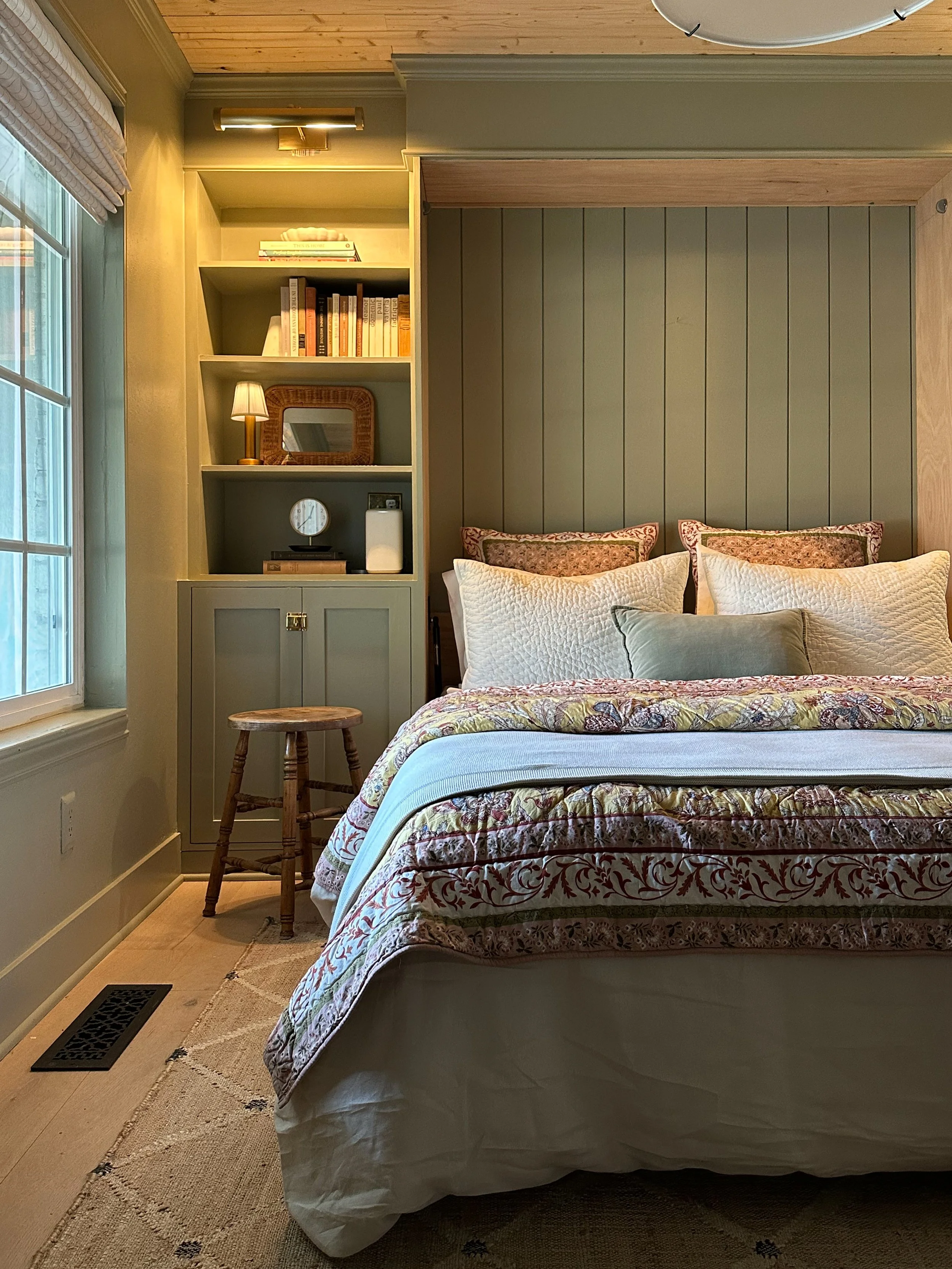 A cozy bedroom with a bed adorned with multiple pillows, a textured bedspread, and a patterned throw blanket. There is a window with a white shade, a lamp on a small wooden side table, and a built-in bookshelf filled with books and decorative items. A framed painting of a landscape hangs above the bed, and the room has a mix of floral and wood paneling wall textures.