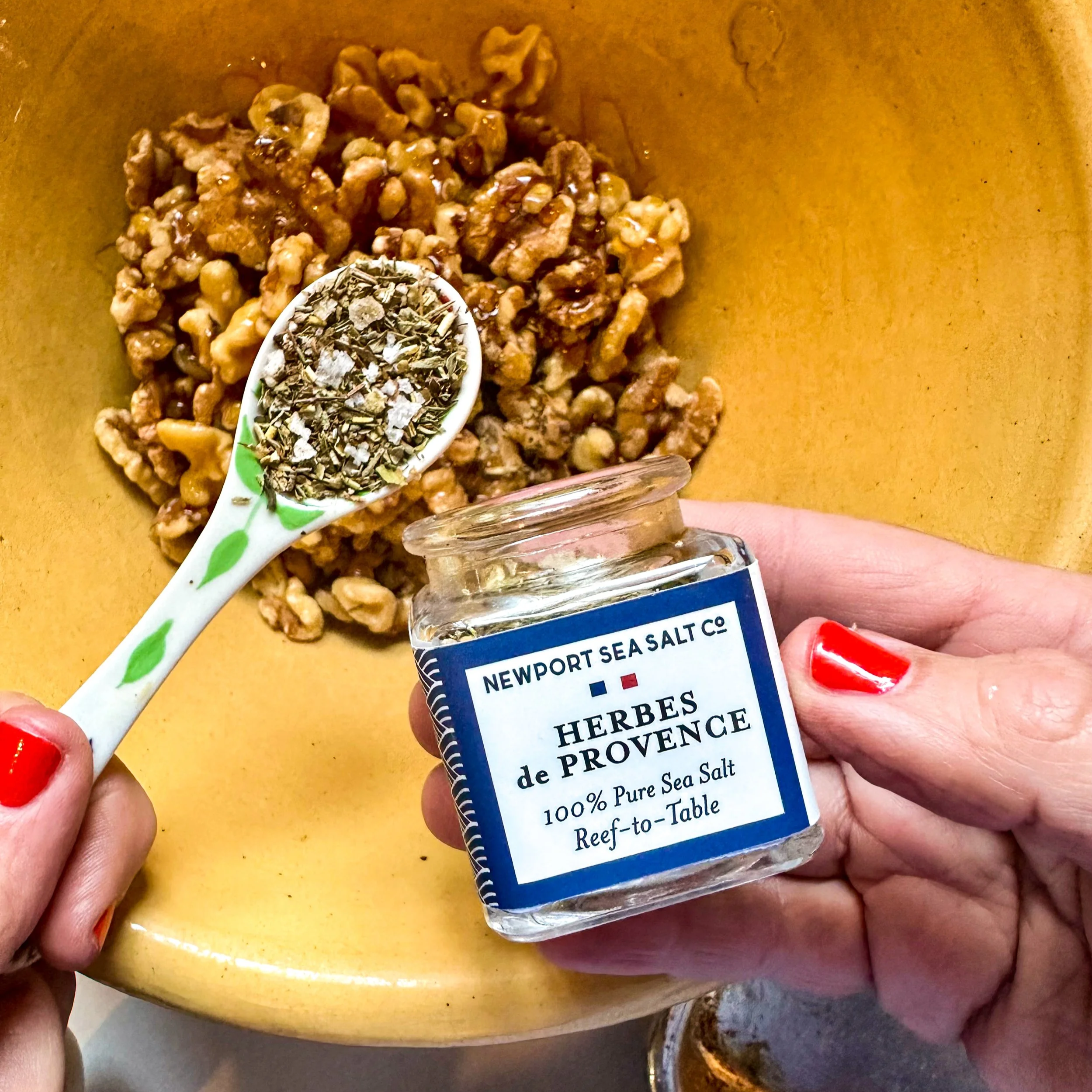 A yellow ceramic bowl full of toasted walnuts pictured with an Herbes de Provence glass jar of seasoned salt along with a teaspoon of the herb blend over the walnuts.