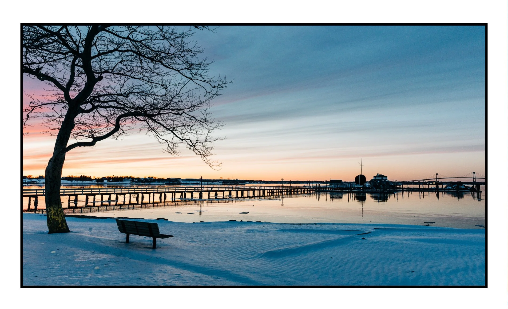 Ida Lewis Yacht Club in the wintertime with a snowy landscape and a lone bench looking off to the Newport Bridge.