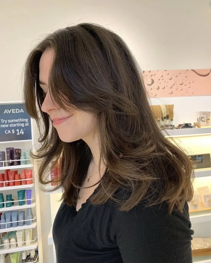 A woman with shoulder-length wavy brown hair and fair skin, smiling softly, in a store with products on shelves in the background.