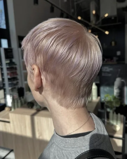 A person with short, layered blonde hair in a modern haircut, seen from the back, in a salon with shelves and lighting fixtures.