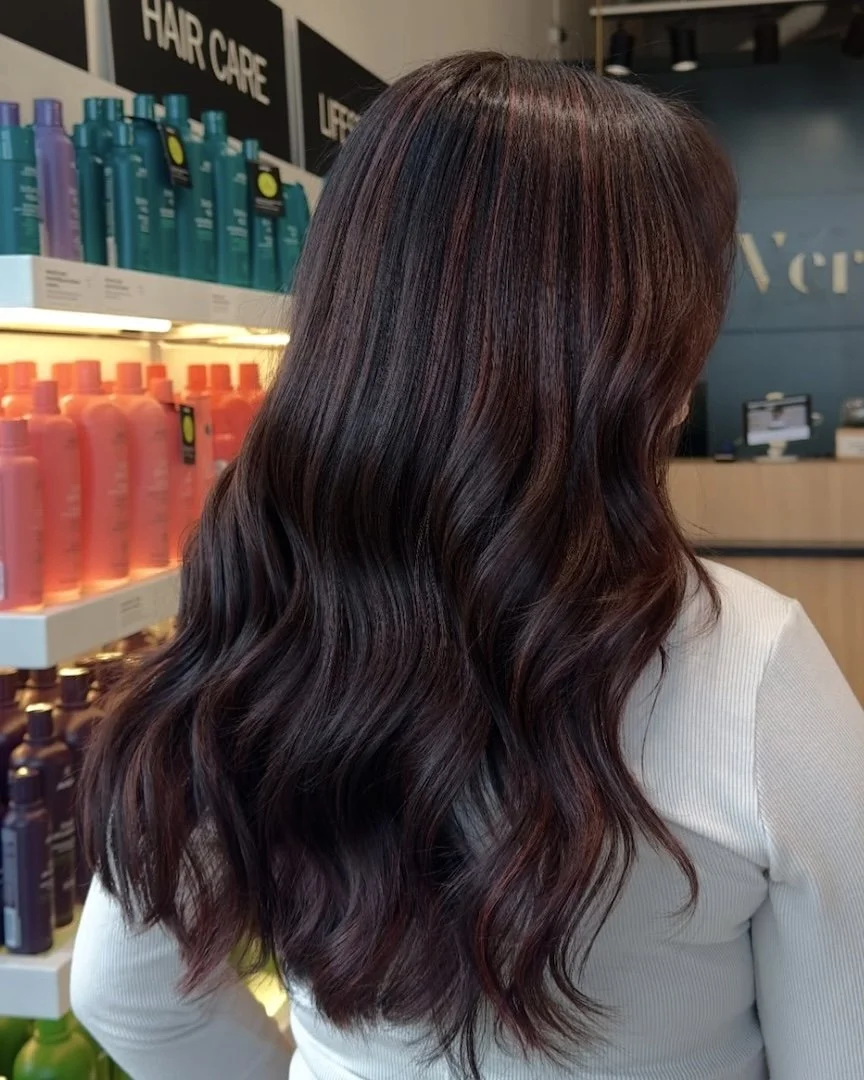 Back view of a woman with medium-length dark hair styled in loose wavy curls, standing in a store aisle with hair care products displayed on shelves.