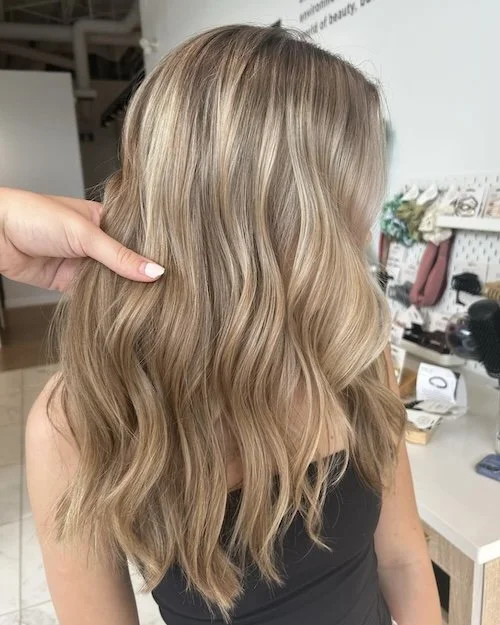 Someone holding a section of long, wavy blonde hair of a woman with her back turned, in a salon environment.