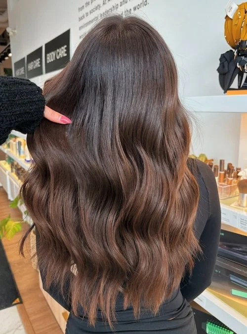 Back view of a woman with long, wavy, brown hair in a store aisle.