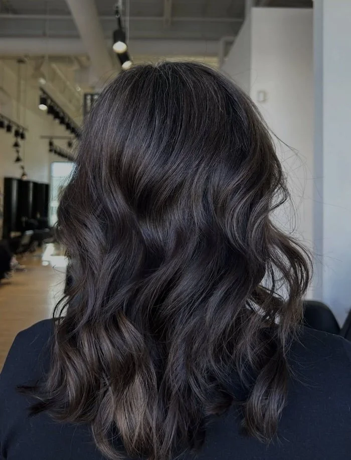 Back view of dark brown, wavy, shoulder-length hair in a salon setting.