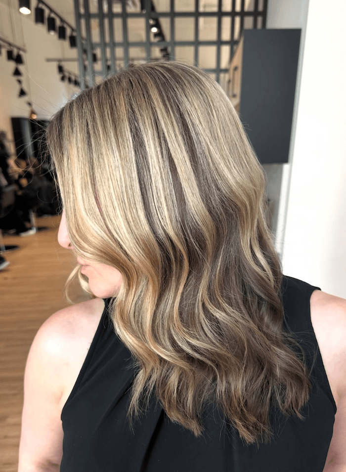 Side view of a woman with long, wavy blonde hair. She is wearing a black sleeveless top and is in a modern salon.