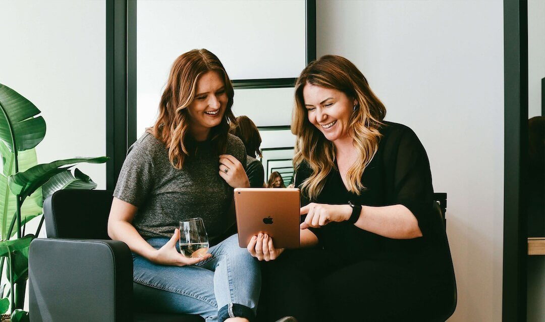 Two women sitting on a dark gray couch, looking at a tablet and smiling, with a glass of white wine in one woman's hand, in a modern office setting with large green plant in the background.