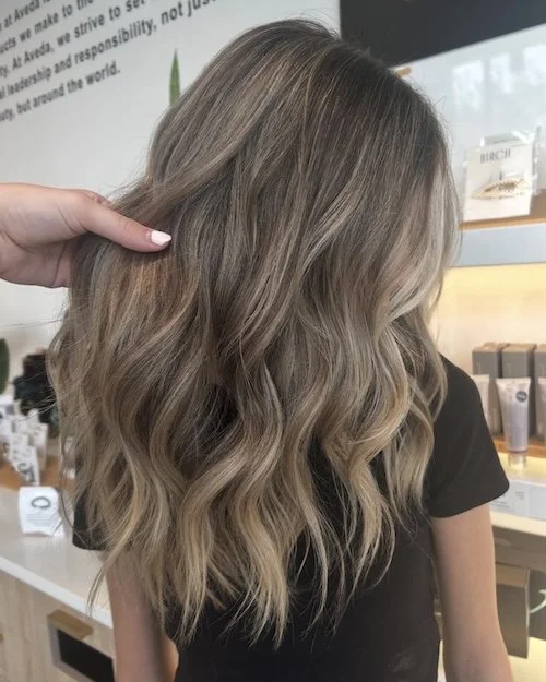 Close-up of a woman with long, wavy, ash-brown hair in a salon, with a stylist's hand gently touching her hair.