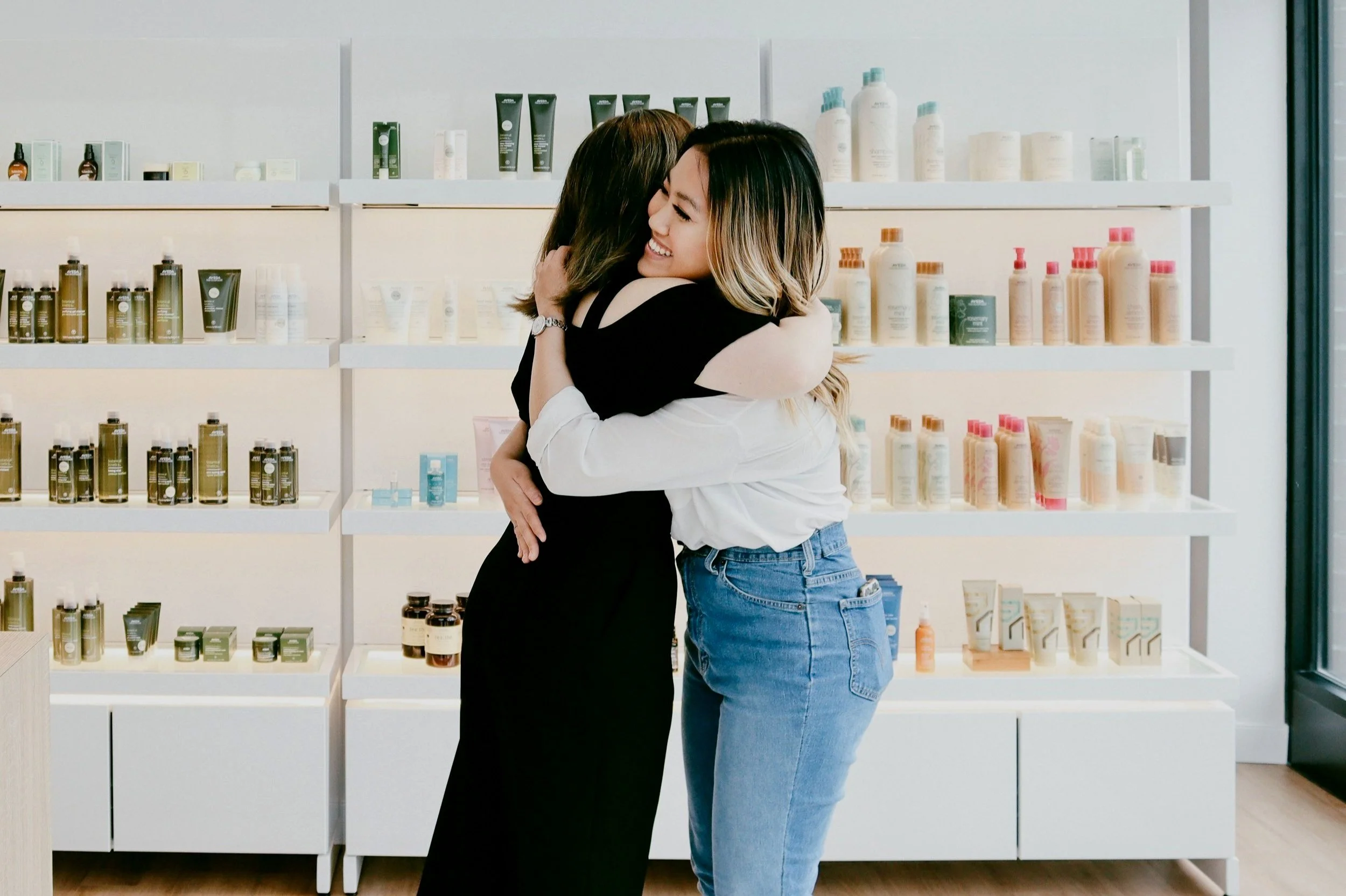 Two women embrace and smile inside a store with shelves of aveda products behind them.