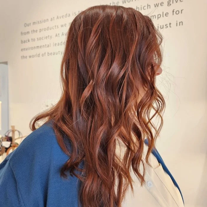 Side view of a woman with long, wavy, reddish-brown hair wearing a beige shirt and dark blue sweater.