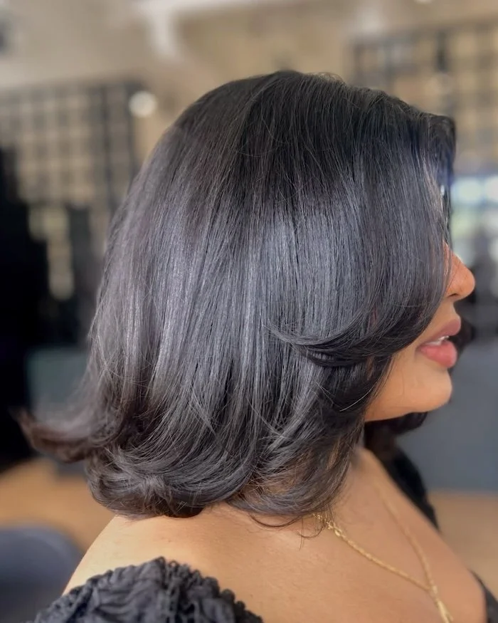 Close-up side view of a woman with a shiny, black bob hairstyle with soft waves, wearing a gold necklace and a black top.