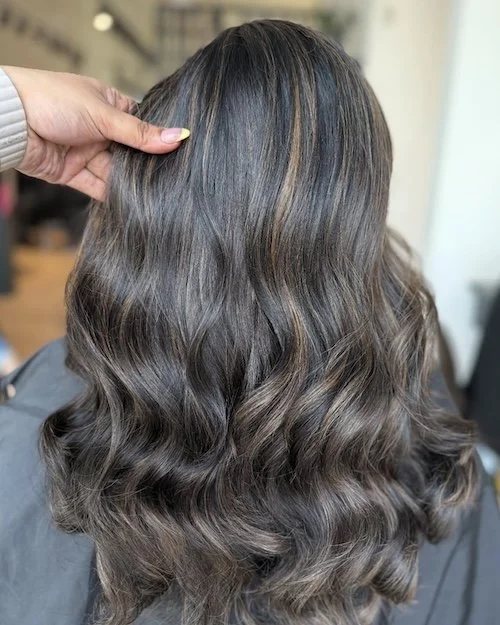 Back of a woman's head showing long, wavy, dark brown hair with subtle highlights, being styled or examined by a person in a salon.