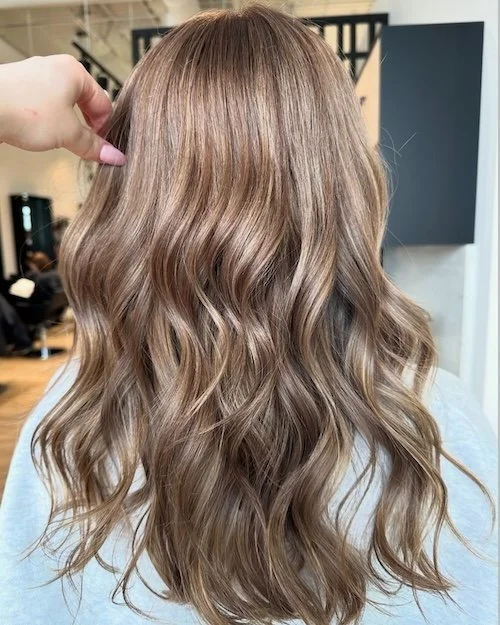 Back view of a person with long, wavy, light brown hair with blonde highlights in a salon setting.