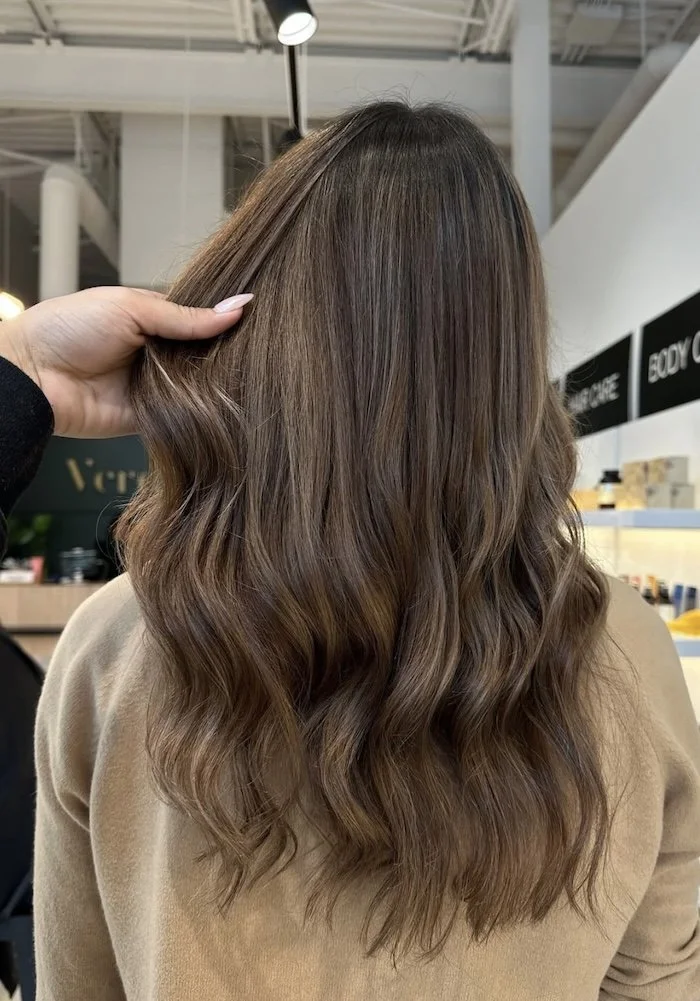 A woman with long, wavy, light brown hair with subtle highlights, seen from behind in a salon or store.
