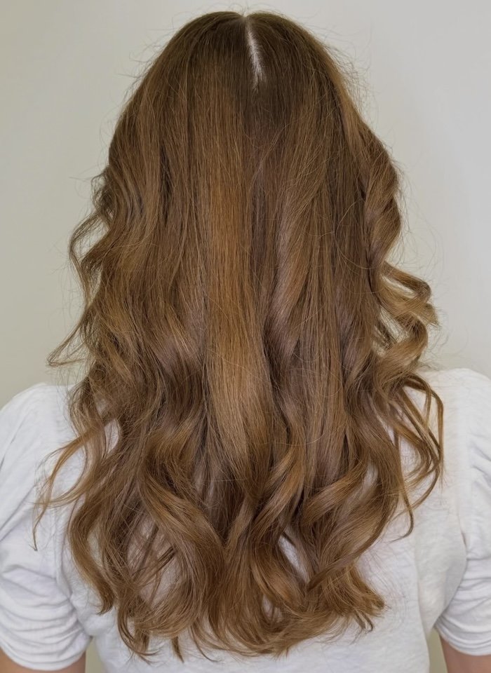 Back view of a woman with long, wavy, reddish-brown hair wearing a white shirt.