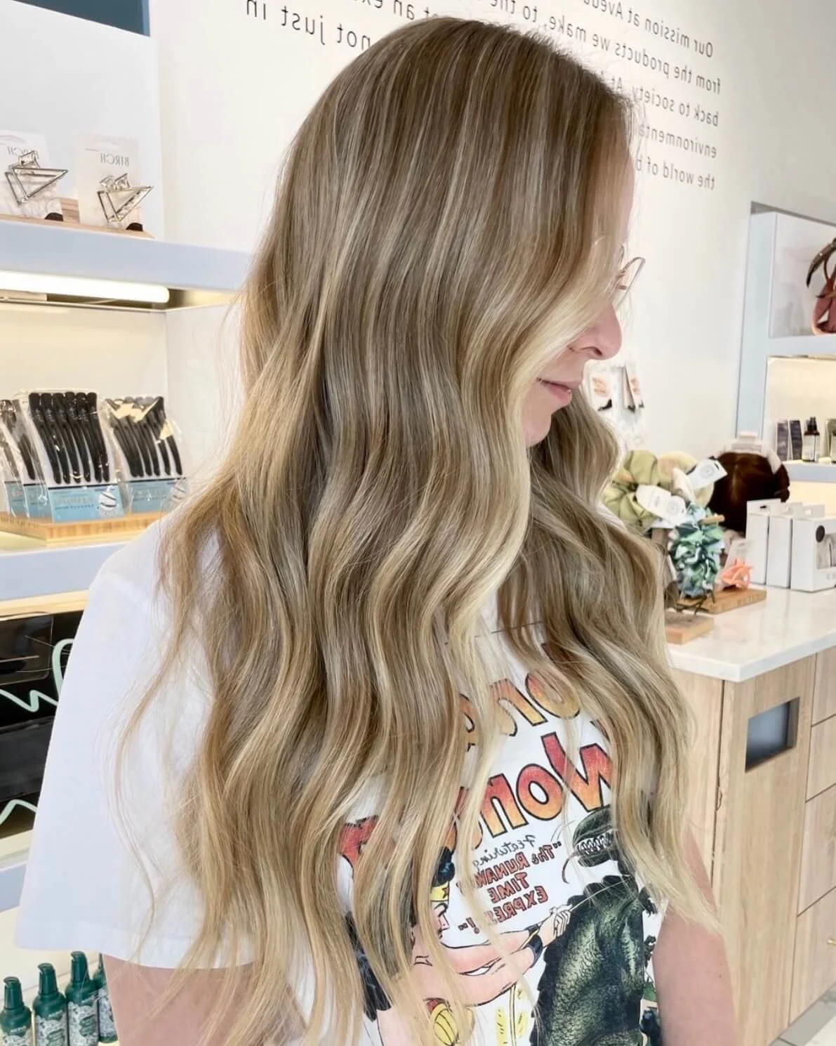 A woman with long, wavy blonde hair standing inside a store, with shelves of hair care products and accessories in the background.