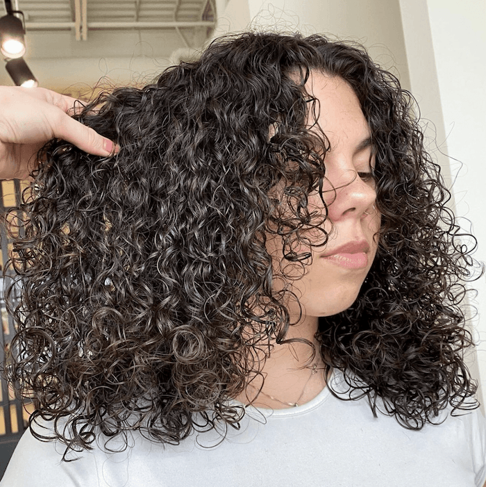 A woman with dark, curly hair is touching her hair with her left hand and looking down.