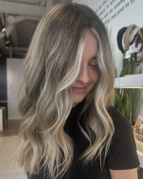 A woman with long, wavy blonde hair and light highlights, smiling with her eyes closed.