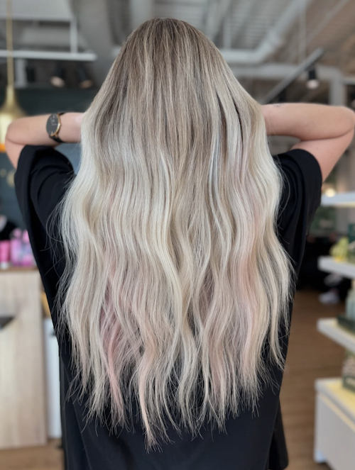 Back view of a woman with long, wavy blonde hair with pink highlights, standing in a modern indoor space.
