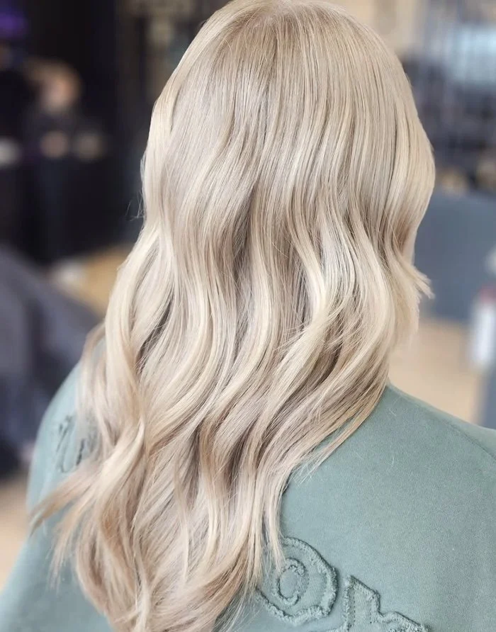 Back view of wavy, blonde hair on a person wearing a green top, indoors with blurred background.