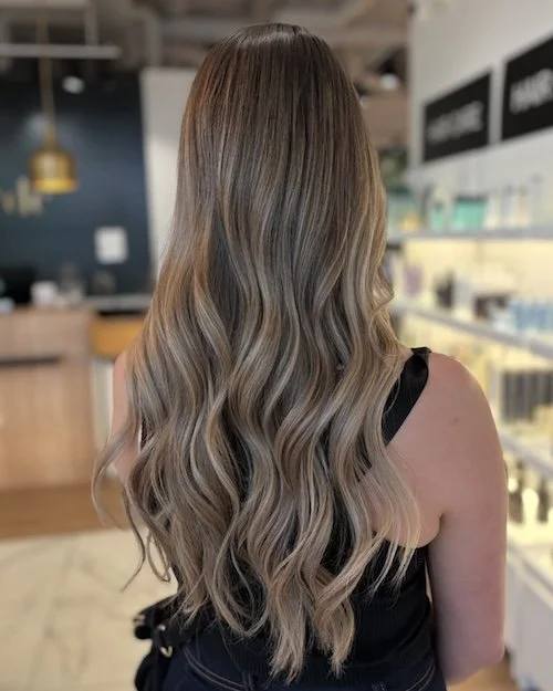 Back view of a woman with long, wavy hair that has a balayage hairstyle in shades of blonde and light brown. She is wearing a black sleeveless top, standing in a retail or salon environment.