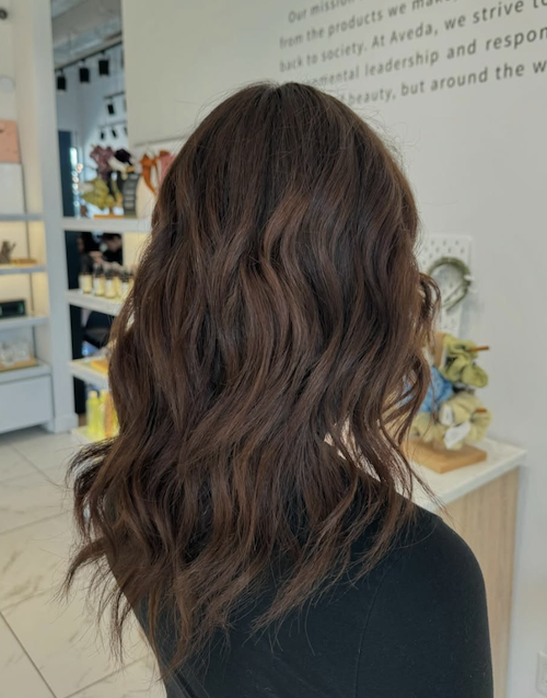 Back of a woman with shoulder-length wavy brown hair, standing inside a store with shelves of products in the background.