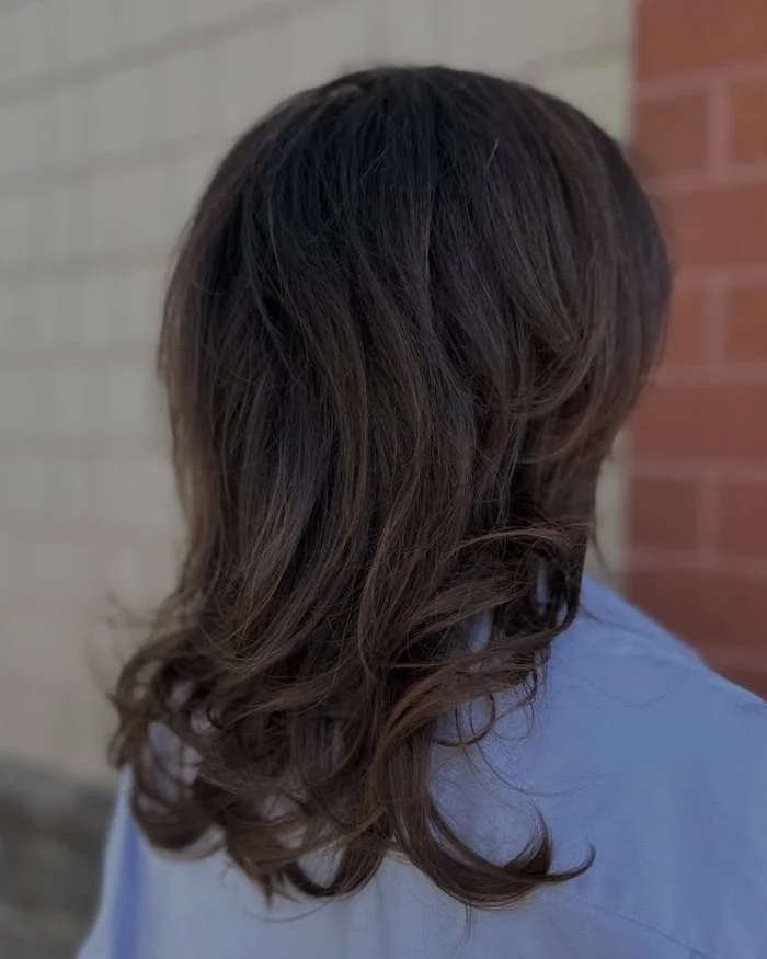 A person with shoulder-length wavy brown hair standing outdoors near a brick wall.