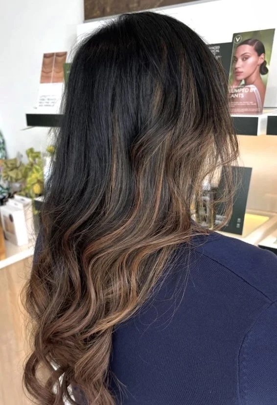 A woman with wavy, shoulder-length brown hair with lighter highlights, sitting in a salon or store. The background includes hair care products and promotional display posters.