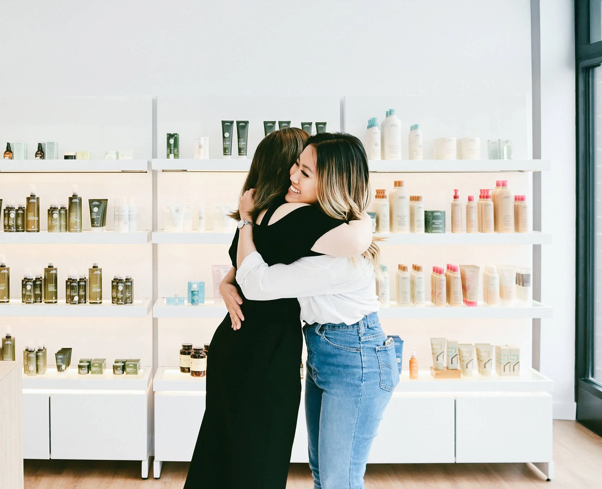 Two women hugging and smiling in front of shelves filled with Aveda products in a bright salon.