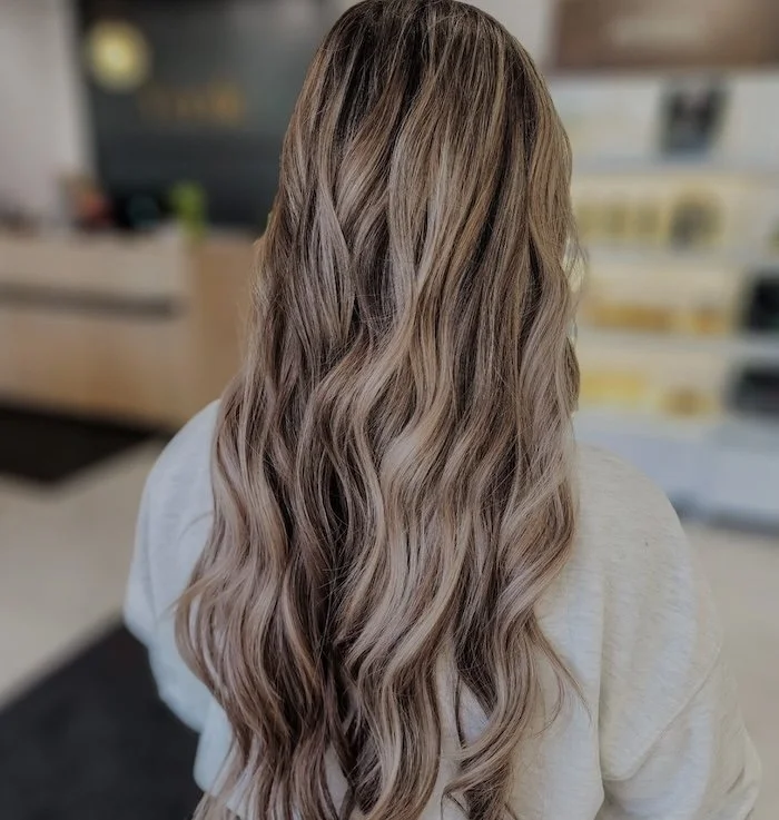 Back view of a woman with long, wavy, light brown hair standing in a room with blurred background.