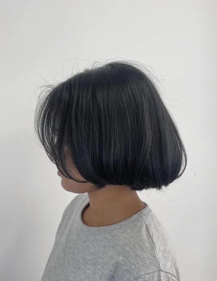 Side view of a person with short, black hair in a bob cut, wearing a light gray shirt against a plain white background.