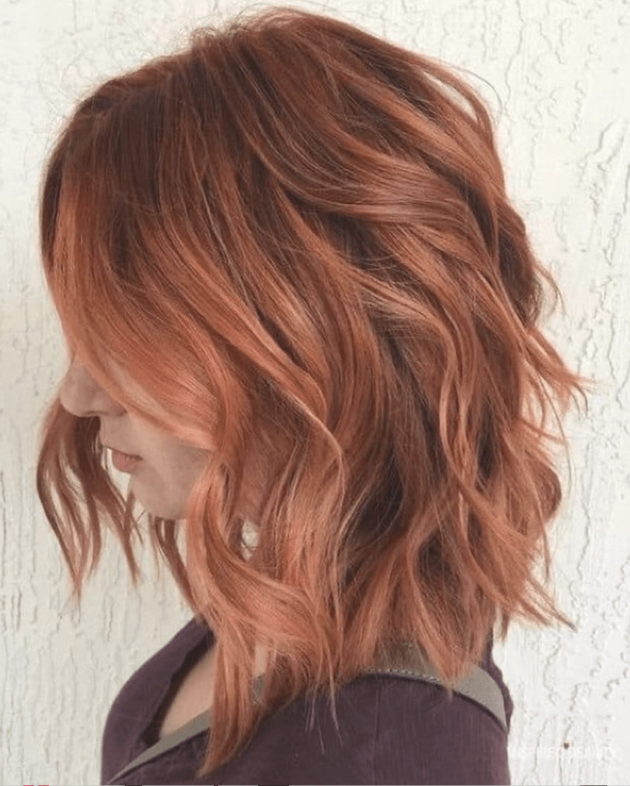 Side view of a woman with shoulder-length wavy red hair against a white textured wall.
