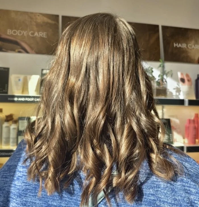 Back view of a woman with shoulder-length, wavy, light brown hair inside a beauty salon.