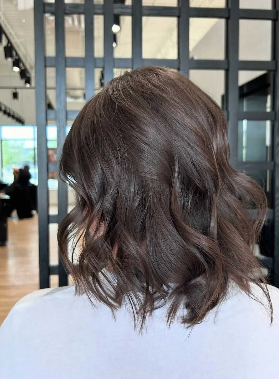 A person with shoulder-length wavy brown hair, seen from behind, in a modern salon with black grid partitions and large windows.