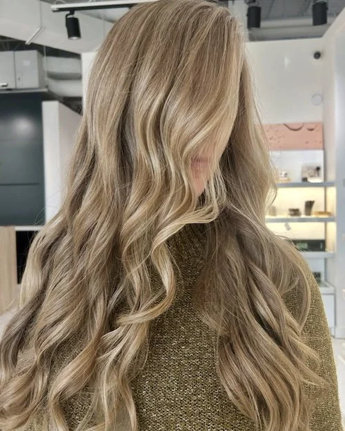 A woman with long, wavy blonde hair, partially covering her face, wearing a brown sweater, in a modern indoor setting.