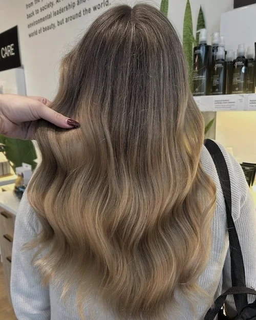 A woman with long, wavy, ombre hair transitioning from dark blonde at the top to light blonde at the ends, in a hair salon. A stylist's hand is holding a section of hair, and salon products can be seen in the background.