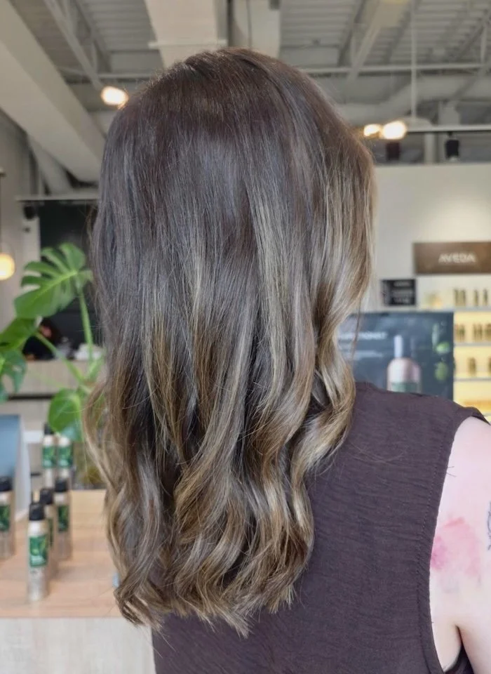 Back view of a woman with medium-length wavy brown hair in a salon.